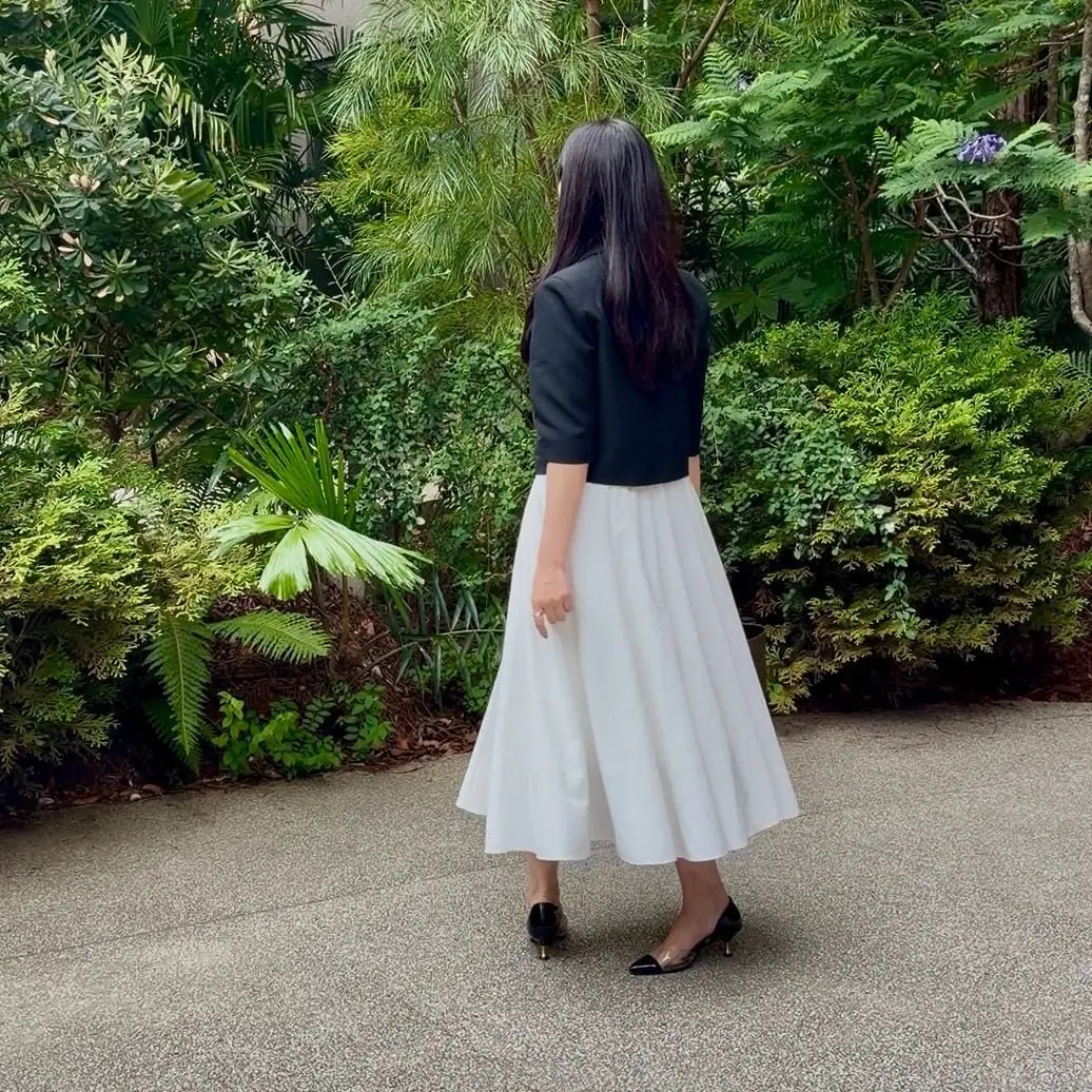 黒いジャケットと白いミモレ丈スカートを着用した女性が、緑豊かな植物の背景に立っている。女性は後ろ姿で、右手をスカートに添えている。足元には黒いパンプス。