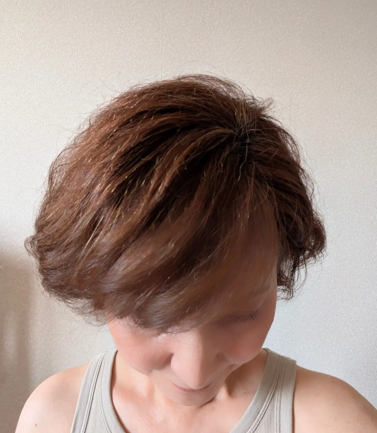 茶色の短髪女性が、無地のグレーのタンクトップを着て正面を見ている。髪は短く、少し乱れたカールがかかっている。背景は白い壁。