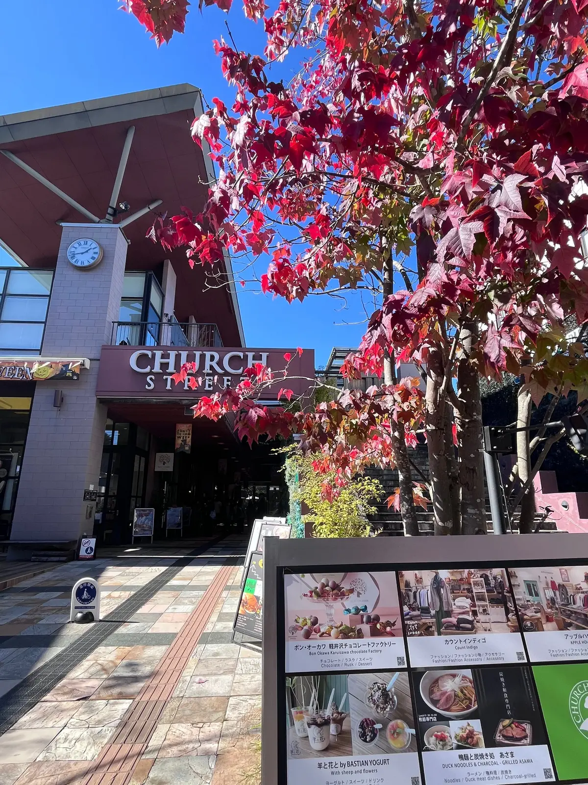秋色の紅葉が茂る並木道。左には「Church Street」と書かれた看板のある建物、正面にはメニュー板が置かれている。背景には時計台が見える。