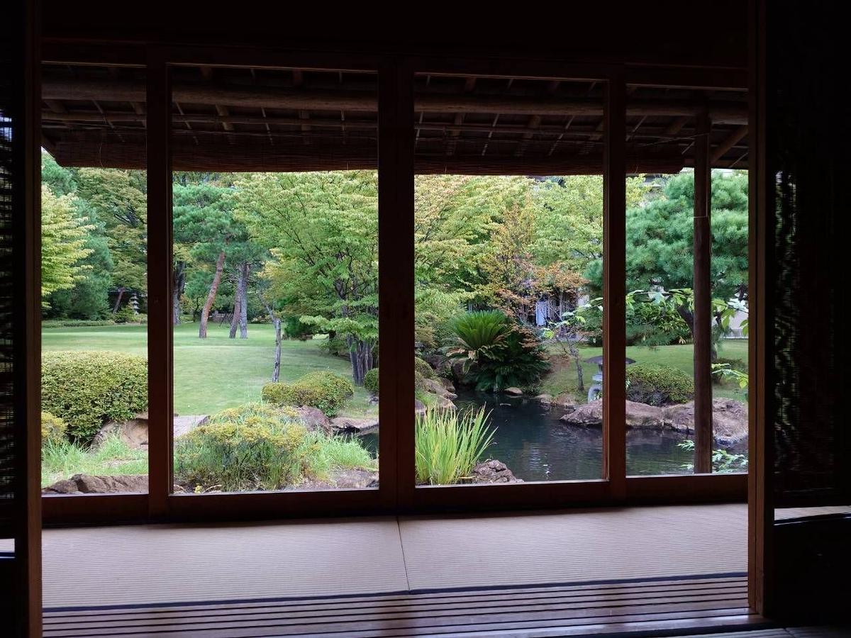 木製の障子を開けた室内から見える、緑豊かな常磐ホテルの日本庭園。手前に池があり、その向こうに草木が生い茂る風景が広がる。庭園は手入れが行き届いており、整然とした美しさがある。