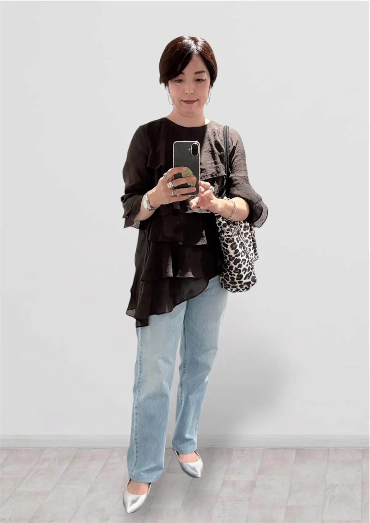 黒い3分袖のトップスにブルーデニムを合わせた女性。右手にスマートフォンを持ち、シルバーのバッグと靴を身につけている。背景は白い壁で、木目調の床。