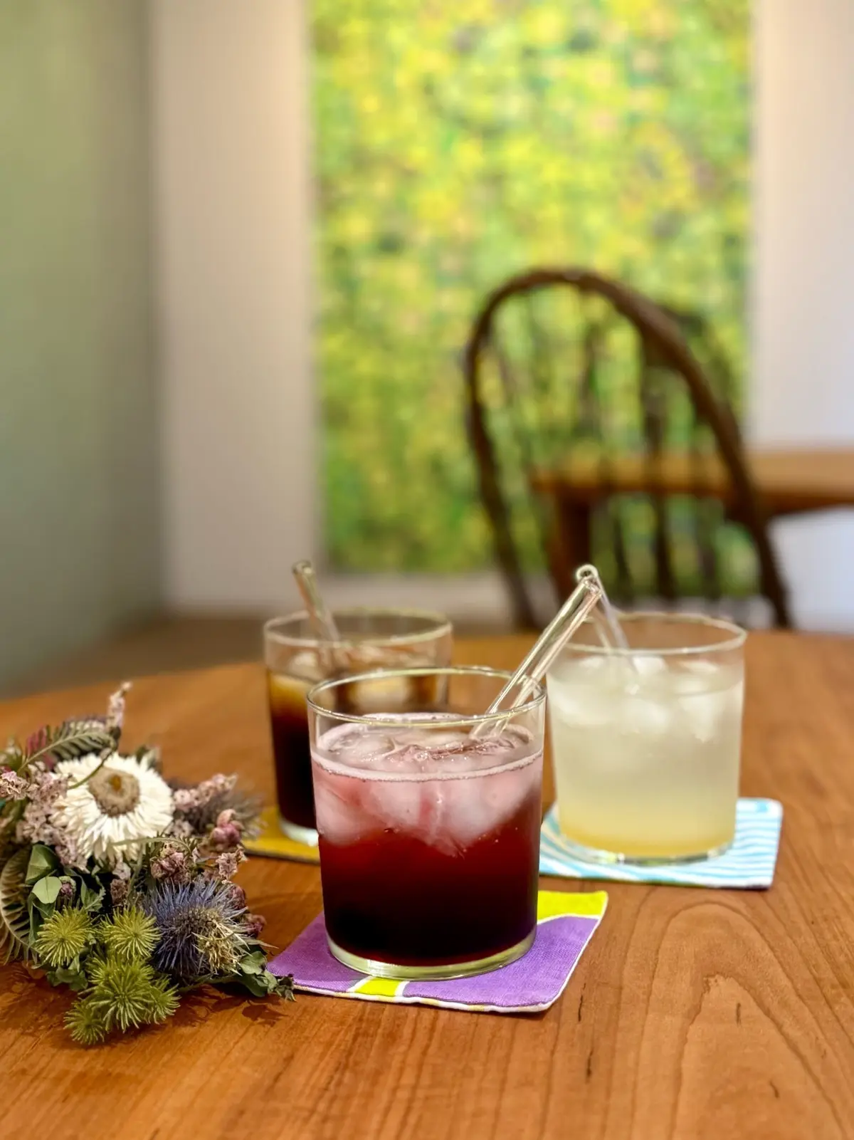 木製のテーブルに置かれた3つのグラス。左からコーヒー、紫色のソーダ、レモネード。それぞれに透明なストローが挿入され、グラスの下にはカラフルなコースターが敷かれている。テーブルの左には花束、背景には緑色の絵画と椅子。
