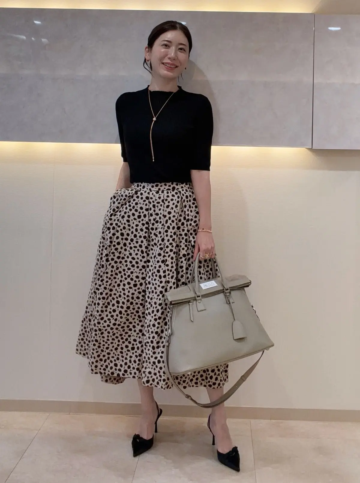 黒のタートルネックに、ベージュのドット柄ミモレ丈スカートを合わせた女性。黒いパンプスを履き、グレーのハンドバッグを持つ。金色のネックレスを着用し、明るい室内で微笑んでいる。
