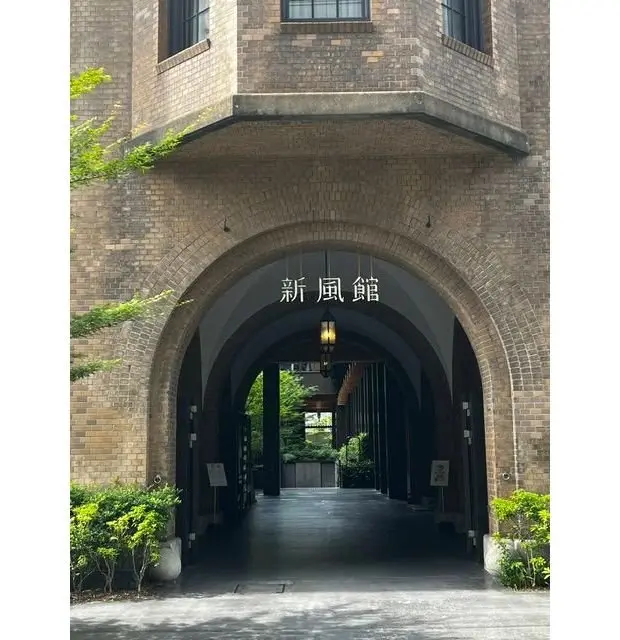 レンガ造りのアーチ門が通路を形成し、中央に「新風館」の文字と照明が設置されている。左右に低木が植えられ、背景に緑の木々が見える。