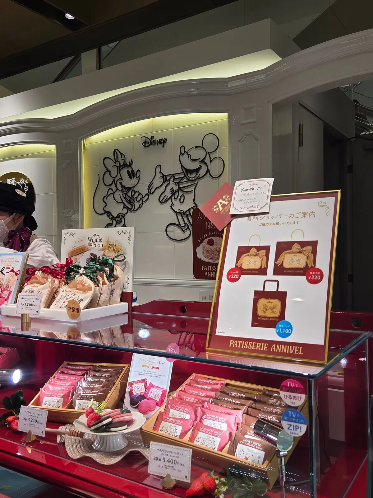 パティスリーアニヴェルヌの店舗外観。窓にミッキーとミニーのイラスト、店内には箱に入ったチョコレートや焼き菓子、イチゴが並ぶ。価格表示やメニューも見える。