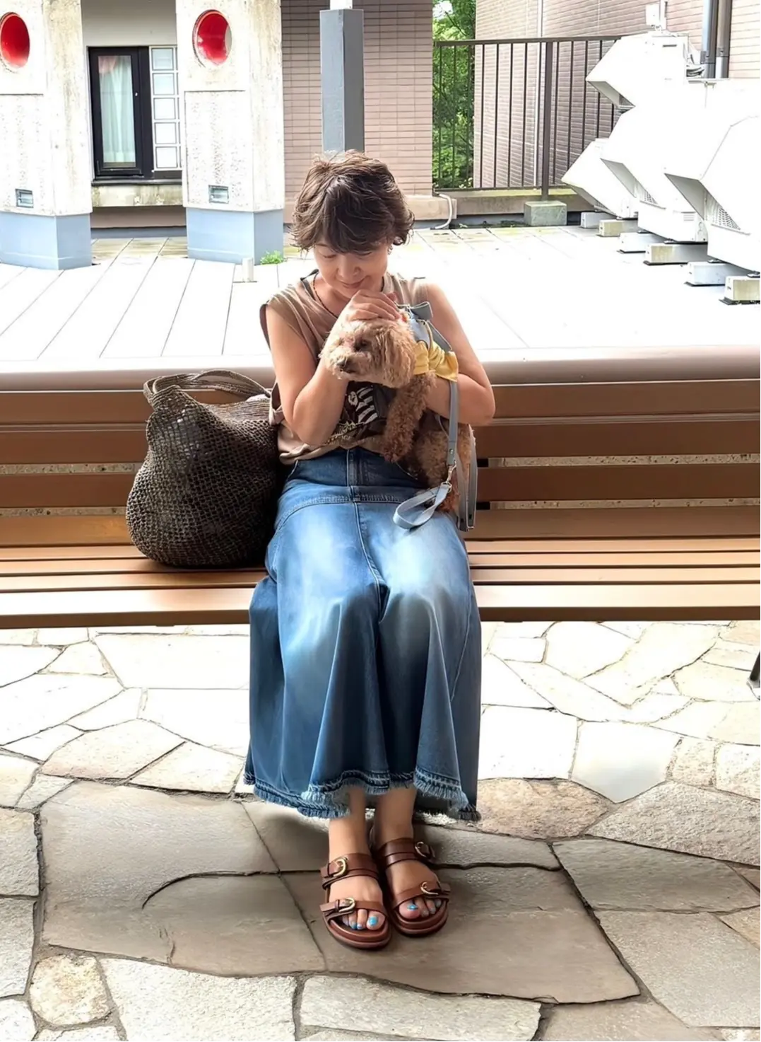 若い女性が、茶色の犬を抱きながら木製のベンチに座っている。女性はベージュのタンクトップにデニムのフレアスカート、茶色のサンダルを着用。大きな編み込みバッグを肩にかけ、背景には白い彫刻がある。