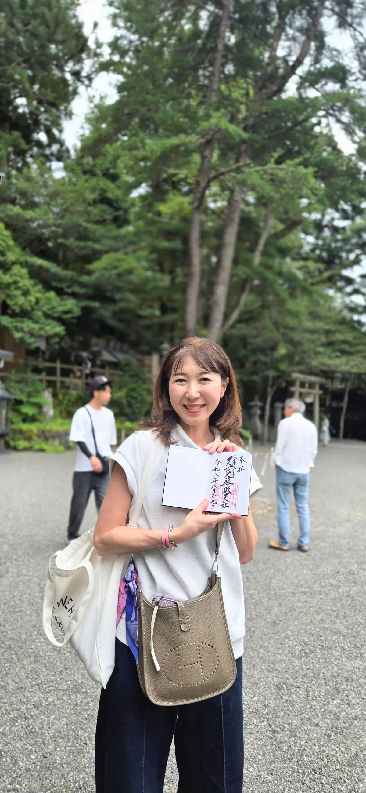 笑顔の女性が、白いシャツにジーンズ姿で屋外に立っている。右手に紙を、左腕にエルメスのバッグをかけている。背景には緑豊かな木々と、人々が歩いている様子が見える。