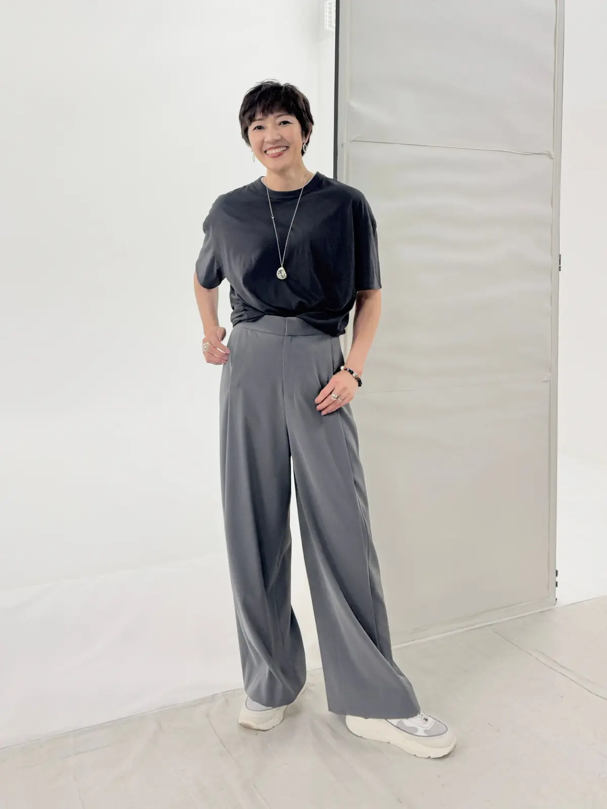 黒のTシャツにグレーのワイドパンツを着用した女性が、白い背景の前で立っている。笑顔でこちらを見ている。シンプルなシルバーのネックレスとリングを身につけている。白いスニーカーを履いている。