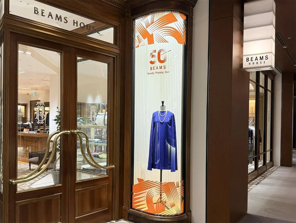 BEAMS HOUSEの店舗入り口。木製の柱と窓枠、大理石調の床タイルが特徴的。右側のショーウィンドウには、青いドレスが展示され、背景にオレンジ色のグラフィックがデザインされている。
