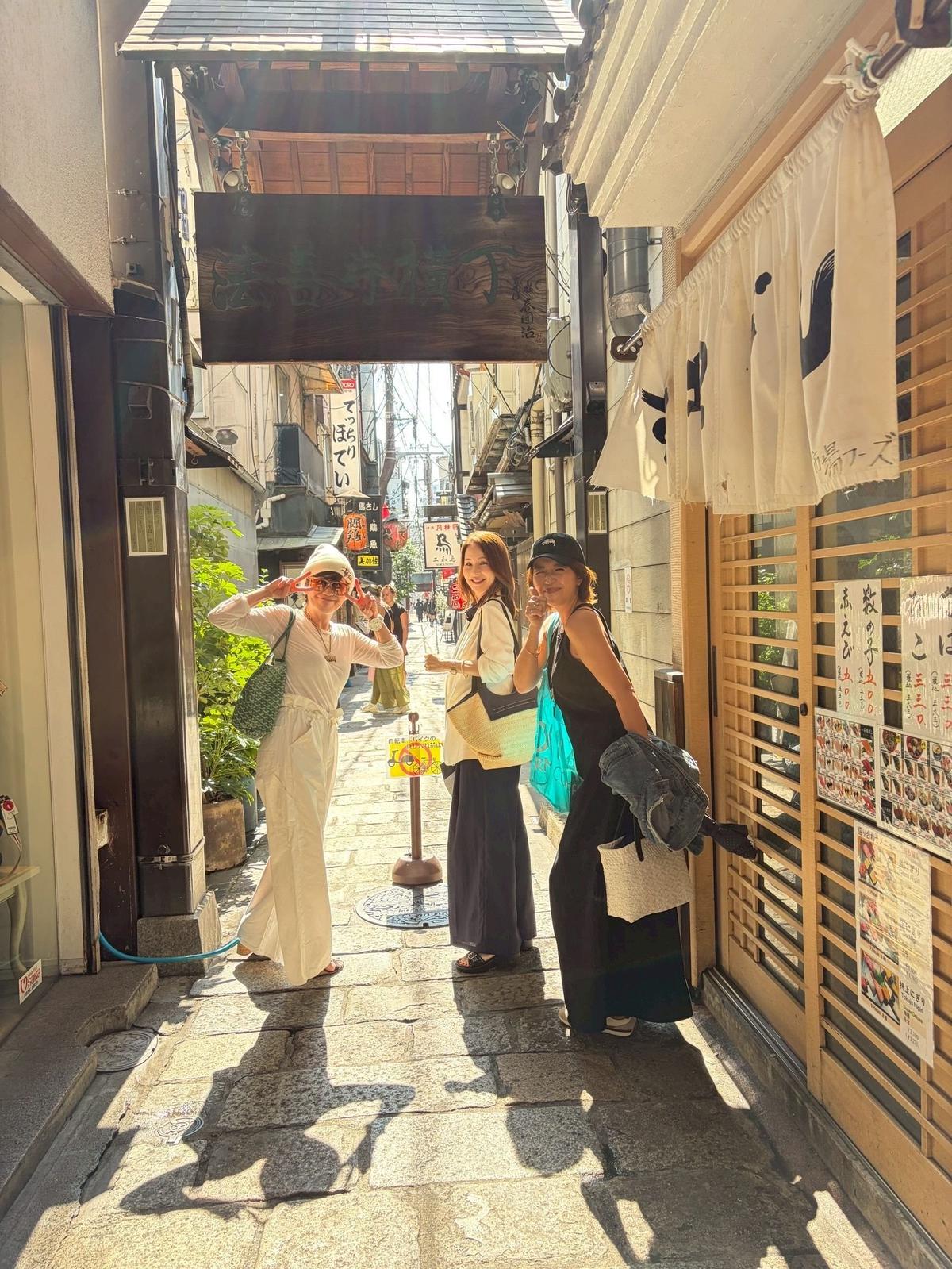伝統的な木造アーチが並ぶ路地で3人の女性が並んで立つ。左の女性は白い服でサングラスをかけ、右の女性は黒い服で帽子をかぶり、それぞれカバンを持っている。背景には店看板や提灯が並ぶ。