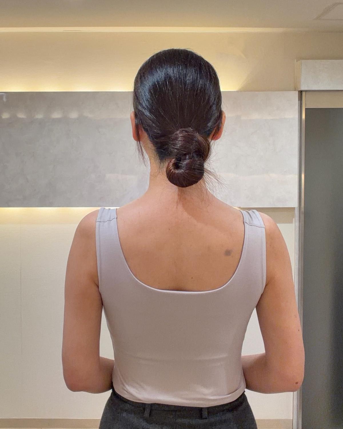 黒髪をまとめ、グレーのタンクトップと黒いズボンを着用した女性の後ろ姿。壁は石調の模様で、シンプルな内装。éclat×BRADELIS ブラタンクトップを着用