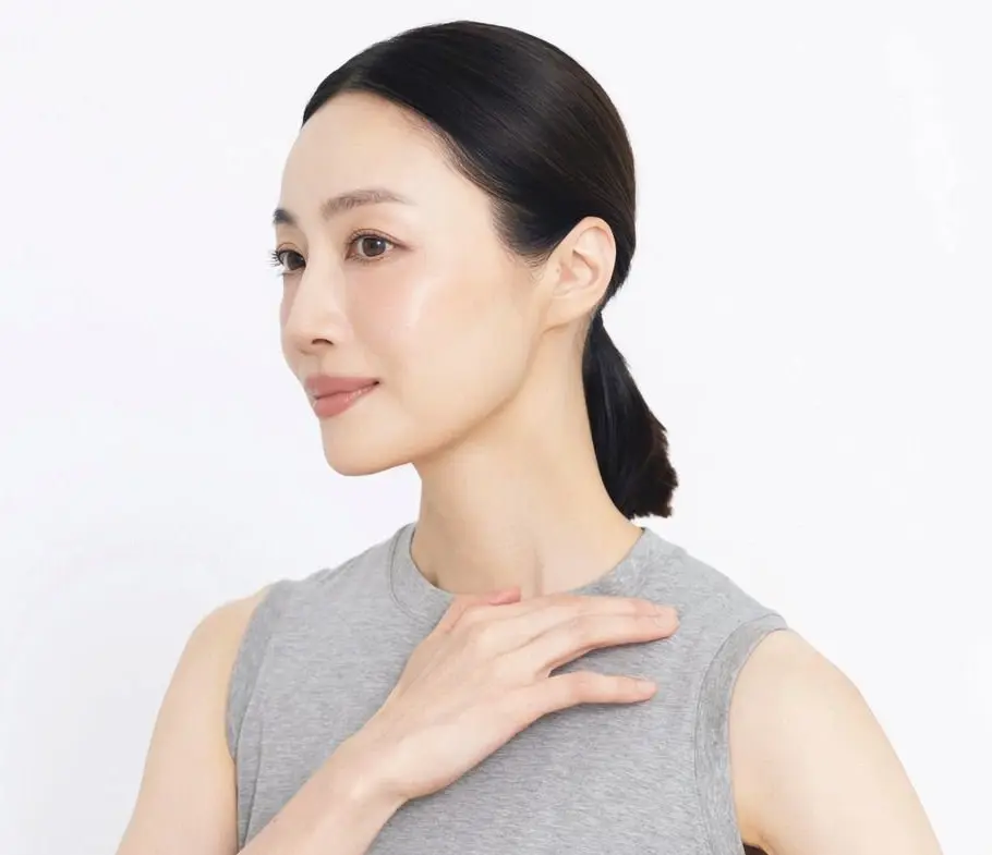 黒髪をポニーテールにした女性がグレーのタンクトップを着て、左手を首に当てています。背景は白です。