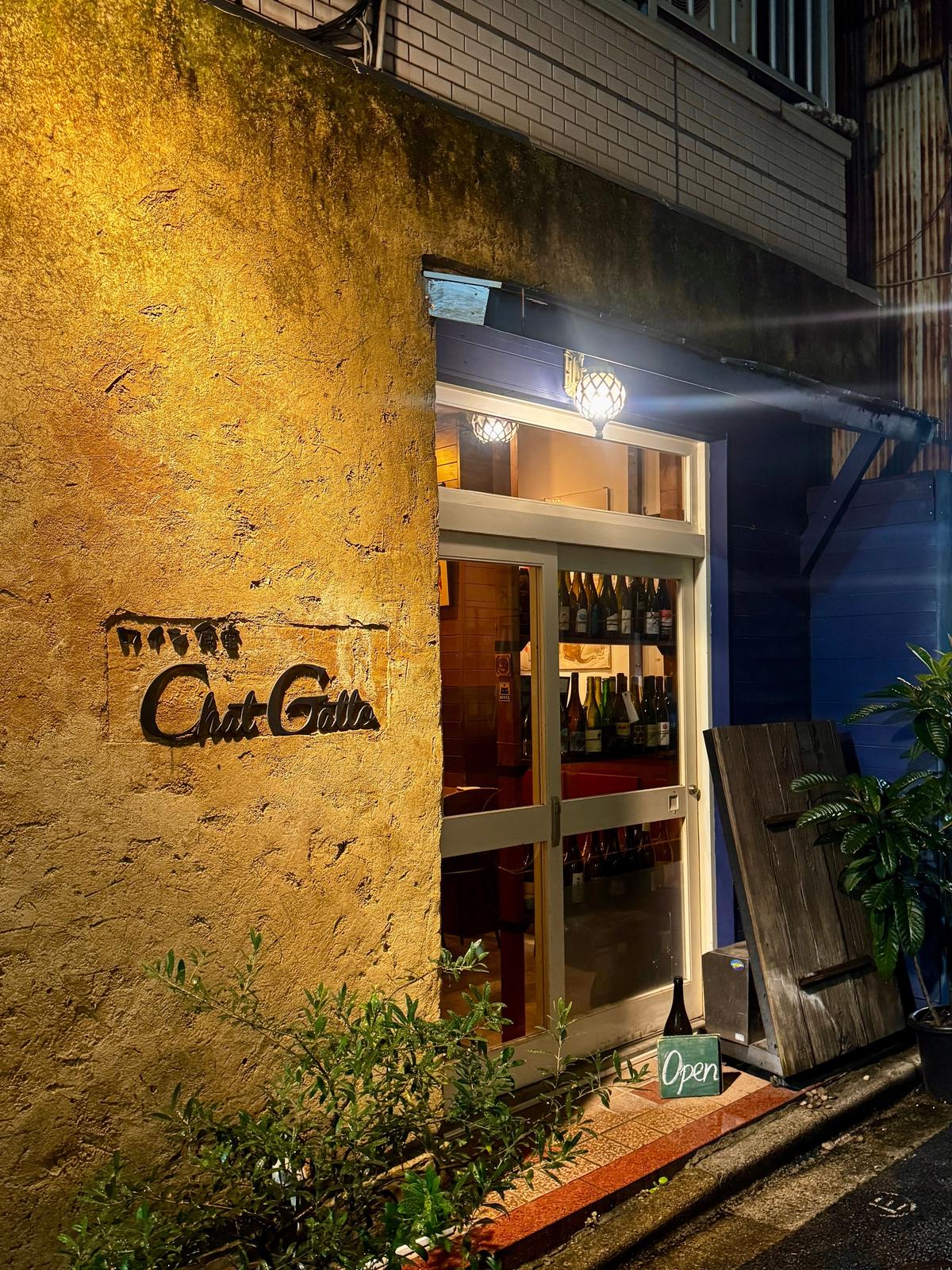 夜の街角に佇む、Chat Galieと書かれた黄色の壁の店。入り口は開いており、内部にはワインボトルが並ぶ。手前に「Open」のサインと植栽が見える。