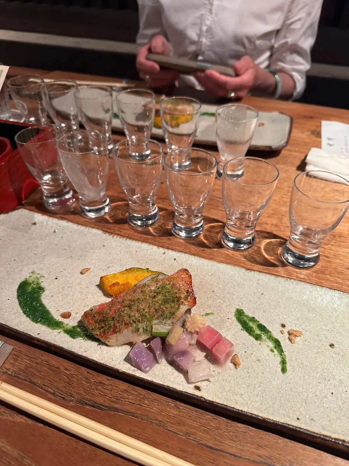 木製のトレイに、緑のソースを添えた魚料理と野菜の盛り合わせ。周囲に複数の空のグラスが並んでいる。