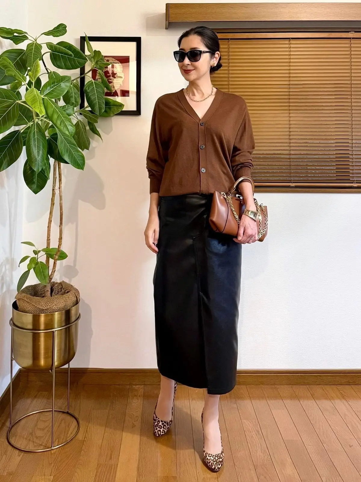 茶色のカーディガン、黒いレザーのミモレ丈スカート、レオパード柄パンプスを合わせた女性。ブラウンのショルダーバッグを斜め掛けし、黒縁サングラスをかけている。背景には観葉植物と窓がある。
GUフェイクレザースカートのコーディネート。