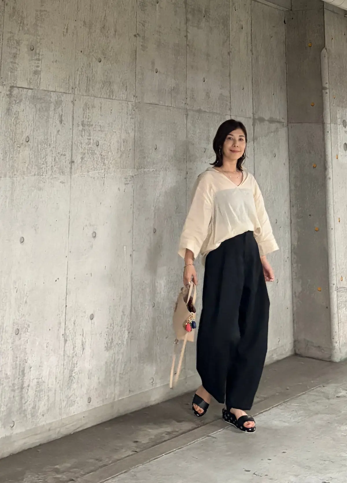 コンクリート壁に寄り添って立つ女性で、ベージュのゆったりした長袖シャツと黒のワイドパンツを着用。右手に小ぶりのトートバッグを持ち、黒のフラットサンダルを履いている。