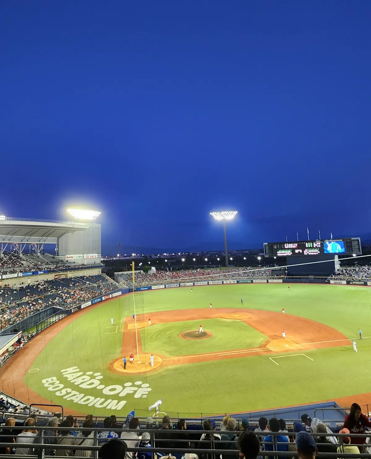野球場に多くの観客が集まり、選手たちがフィールドでプレーしている。夜の照明が明るく、スコアボードが設置されている。フィールド中央には「HARD OFF」という広告が書かれている。