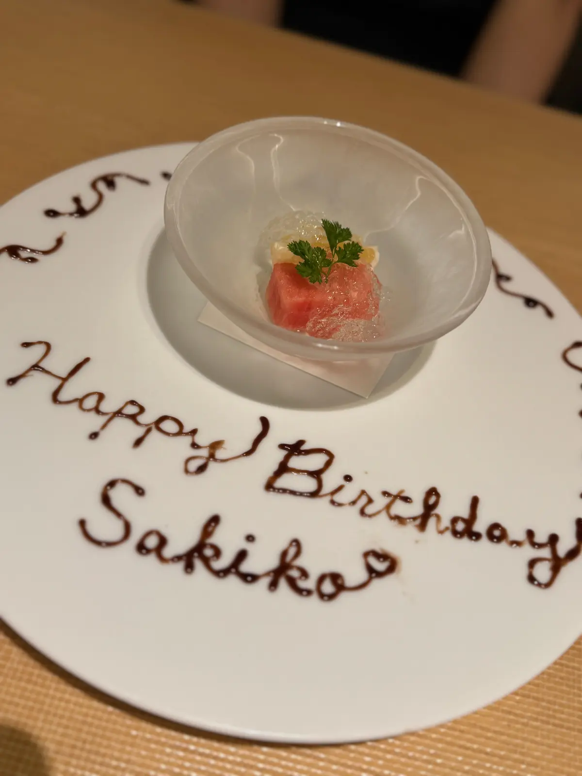 白いプレートに「Happy Birthday Sakiko」とチョコレートで書かれた文字があり、その上に透明なボウルが置かれています。ボウルにはピンク色の肉とパセリが添えられた料理が入っています。