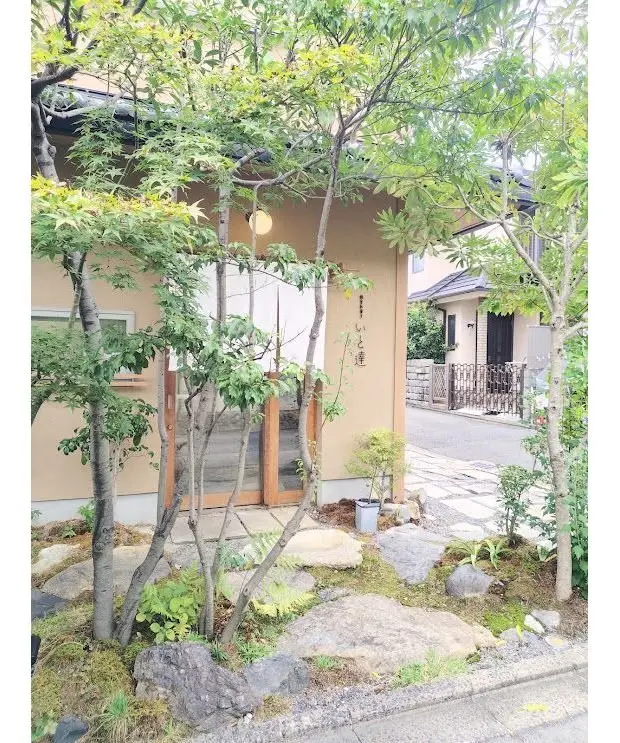 木々が生い茂る庭の前に佇む、ベージュ色の建物の入り口。木製の格子戸が設置され、右側に日本語の文字が書かれた看板がある。石畳の道が続き、背景には別の家が見える。