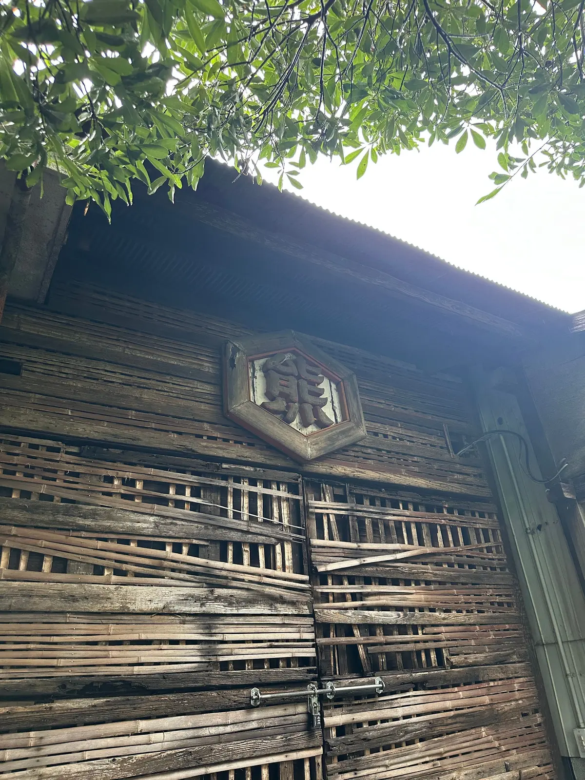 木造の古い建物の壁、竹製の格子と木製の扉、屋根の上に緑の葉が見える