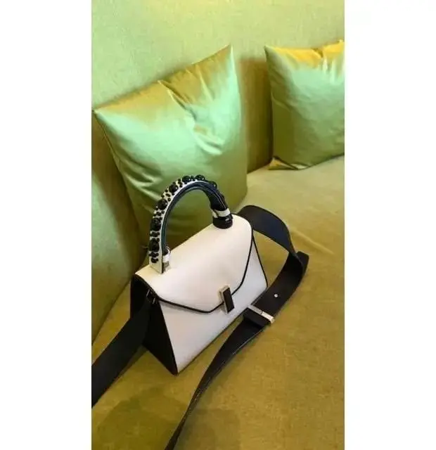 オフホワイトのショルダーバッグ。黒のトリミングとハンドル、シルバーのバックルがアクセント。背景には黄緑色のクッションが置かれている。