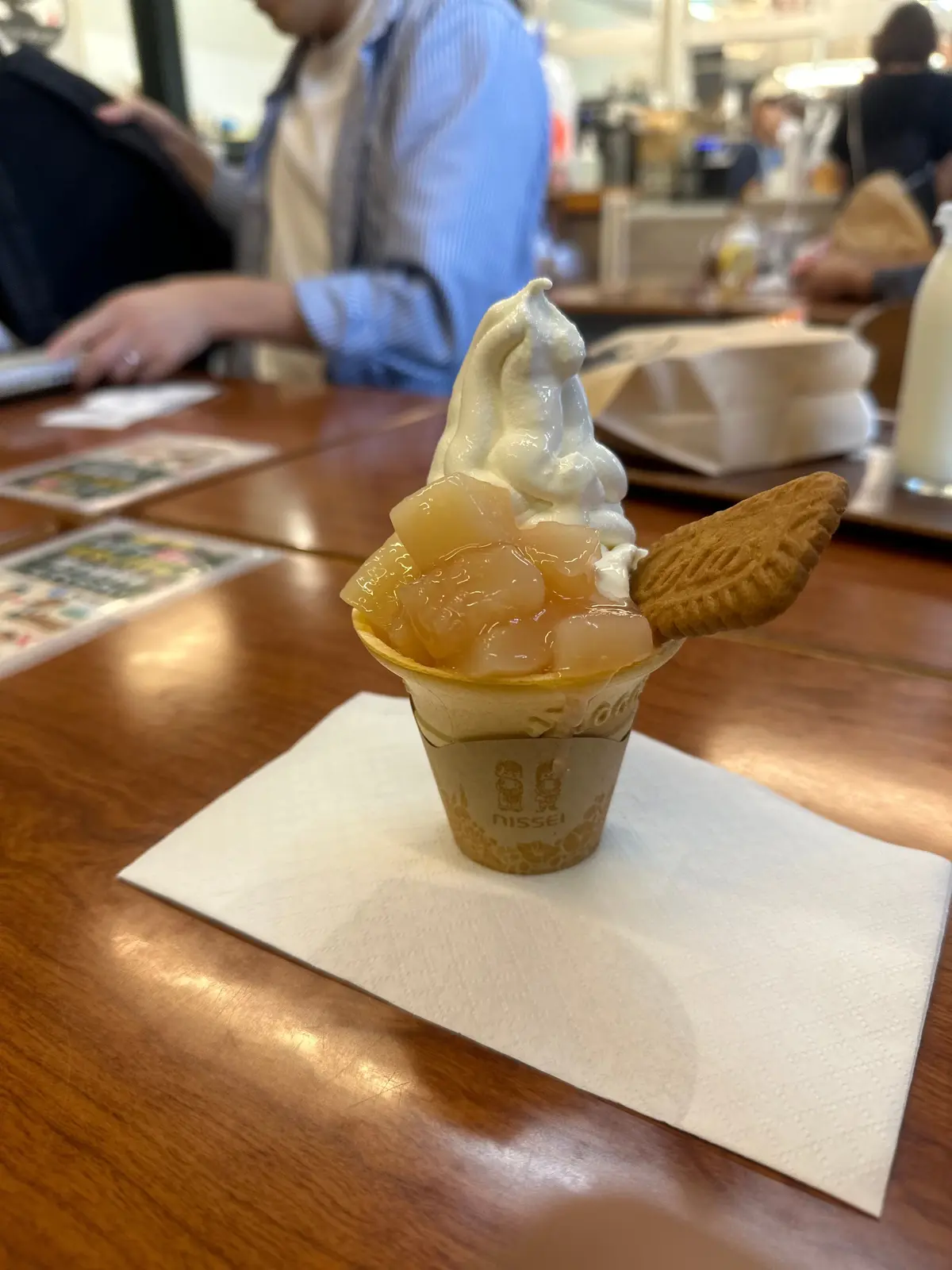 木製のテーブルに置かれた、ソフトクリームのアップ。バニラ色のソフトクリームが３つ盛りになり、クッキーとシロップが添えられている。カップには「NISSEI」のロゴが入っている。背景には人物がぼやけて写っている。