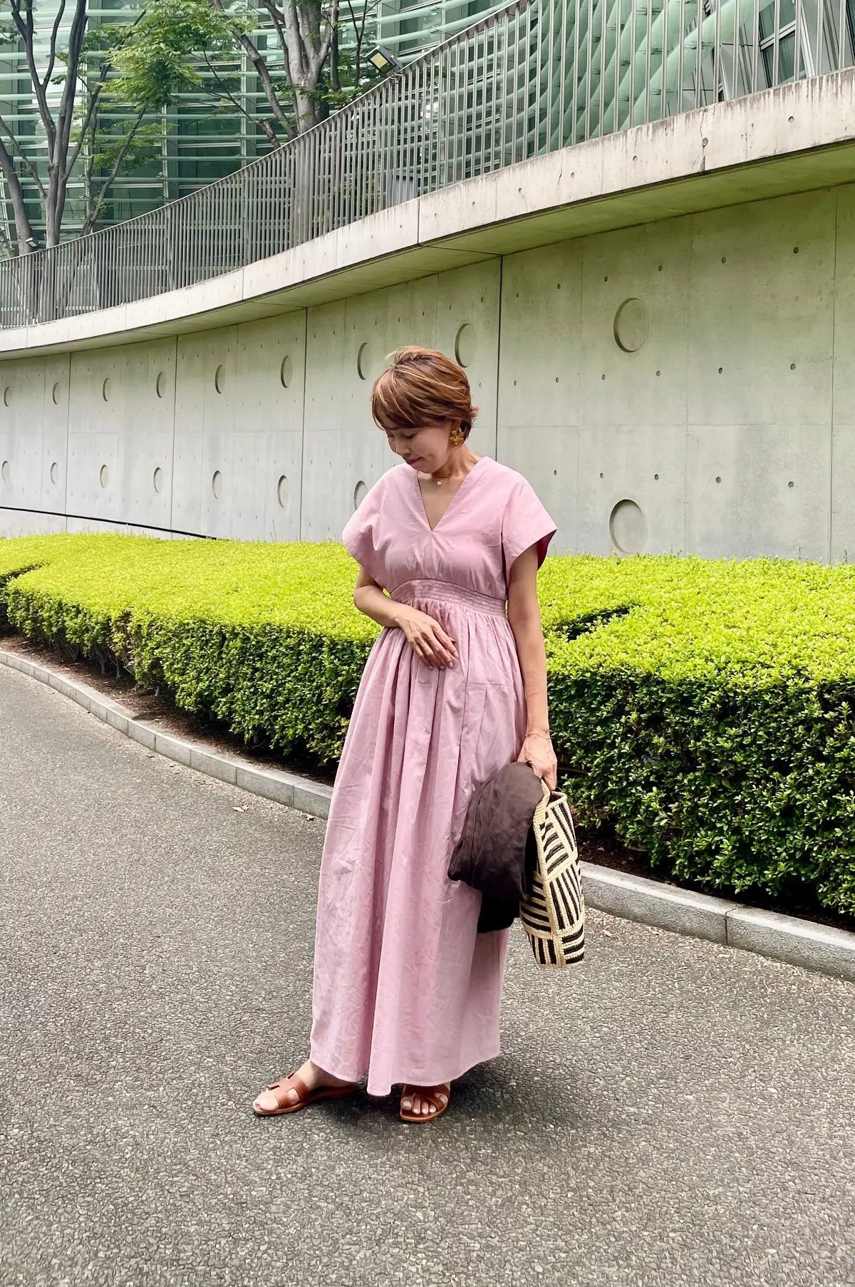 ピンクのロングワンピースを着た女性が、コンクリート壁と低木の並ぶ歩道を歩いている。女性は手袋とストライプ模様のトートバッグを持っている。背景にはガラス張りの建物と木々が見える。