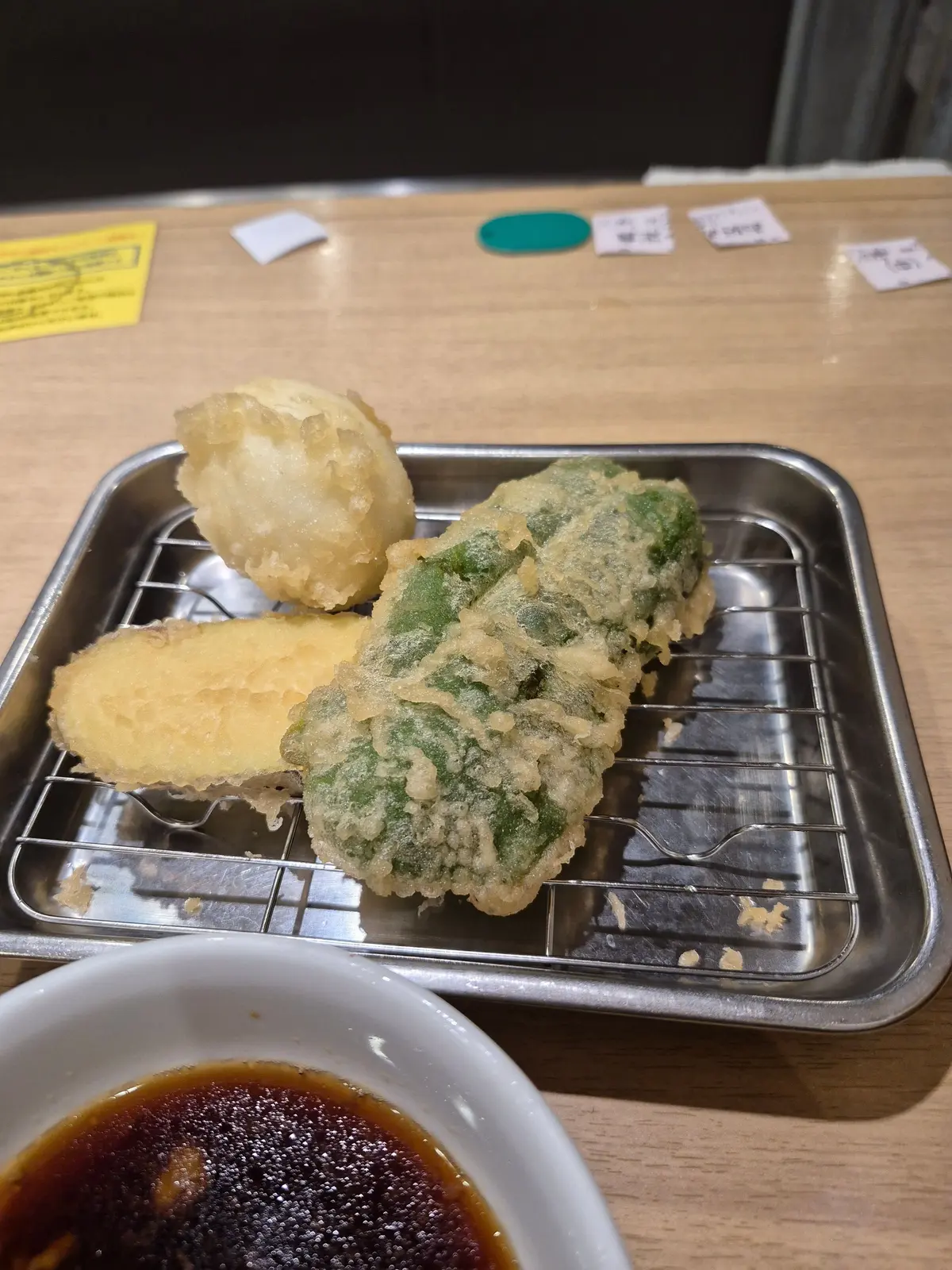 金属製のトレイに盛り付けられた、３種類の天ぷらが置かれている。左から、白身魚、ナス、青菜の天ぷら。小皿の脇には、醤油だれが入った白い器が置かれている。
