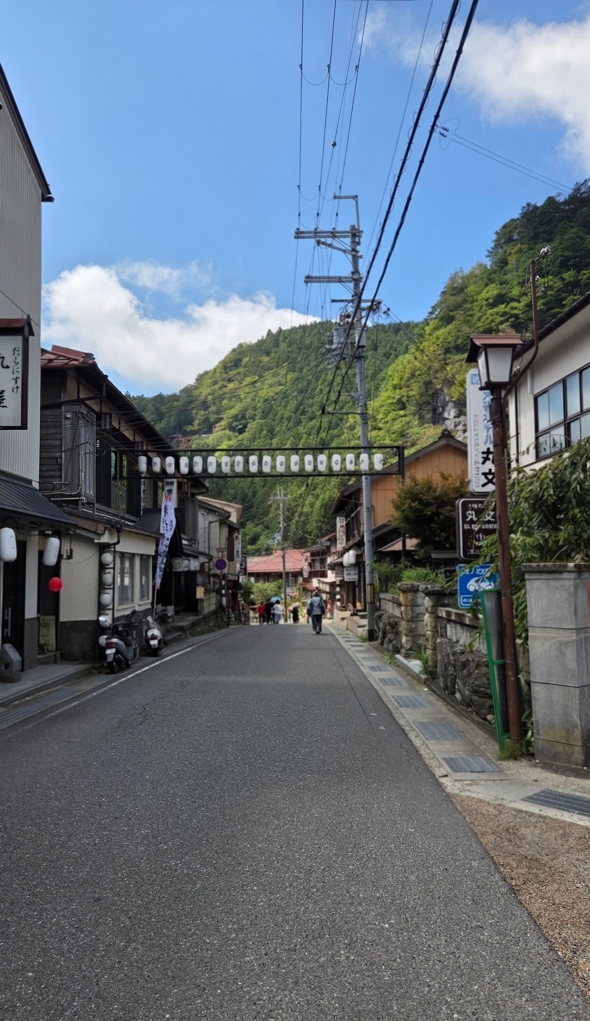 山あいの街並み。細い道が山に向かって続き、左右には木造の建物が並ぶ。電線が張り、遠景には緑の山々が広がる。
