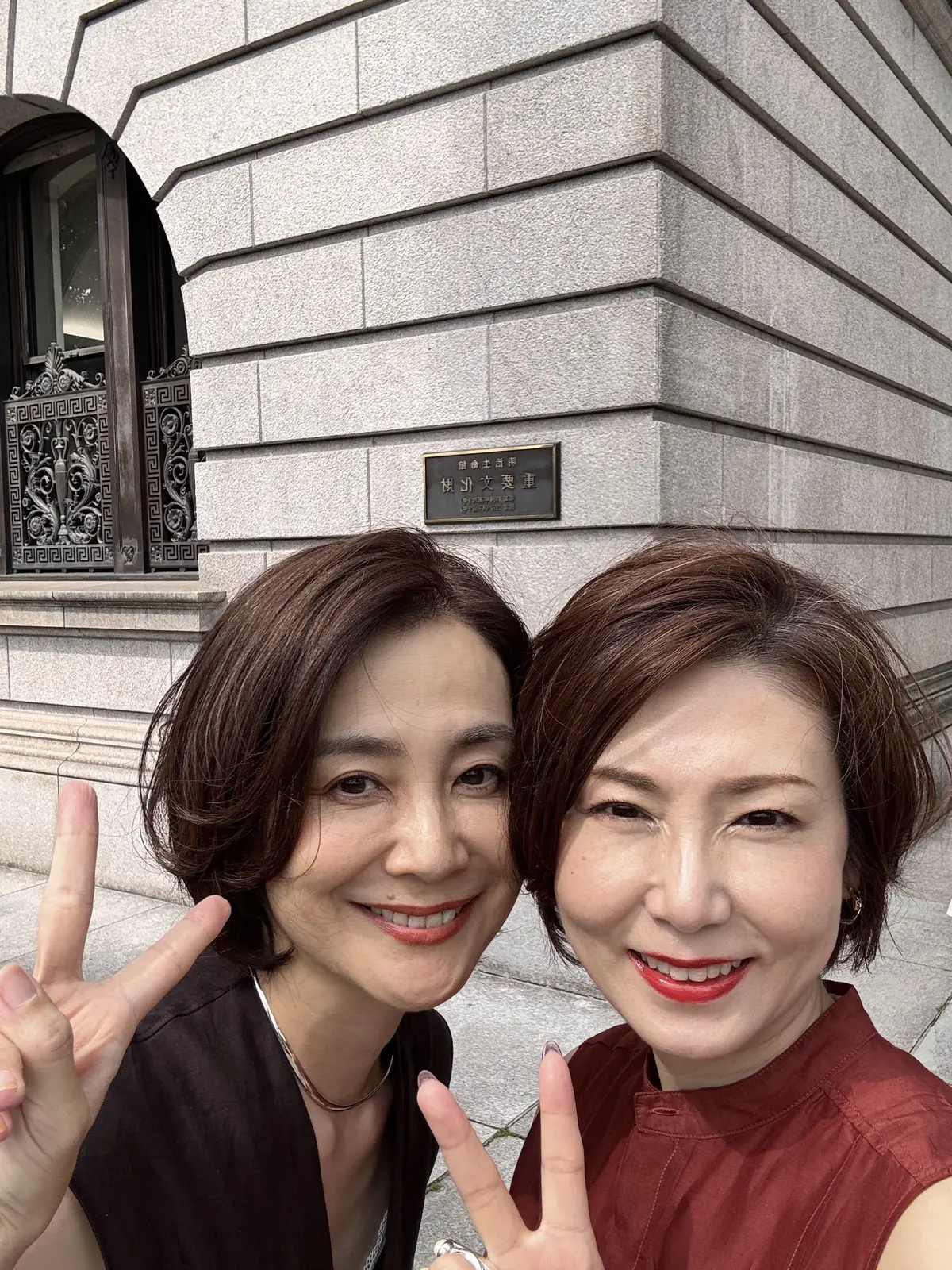 2人の女性が笑顔でピースサインをしている自撮り写真。背景には灰色の石造りの建物があり、黒い装飾の扉と銘板が見える。