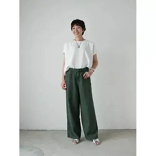 白いTシャツに緑色のワイドパンツを着用した女性が立っている全身写真。女性は微笑んでおり、シルバーのサンダルを履いている。背景は無地で、シンプルなスタジオのような雰囲気。