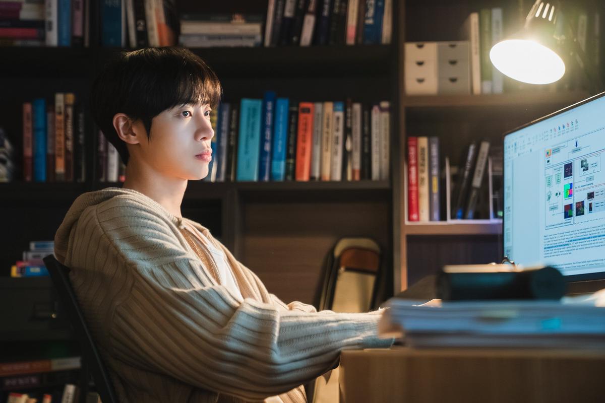 韓国ドラマ　Disney+ (ディズニープラス)　『恋愛の博士課程』 机に向かってパソコンの画面を見つめる若い男性。背景には本棚があり、デスクライトが光を当てている。白いパーカーを着て、真剣な表情をしている。