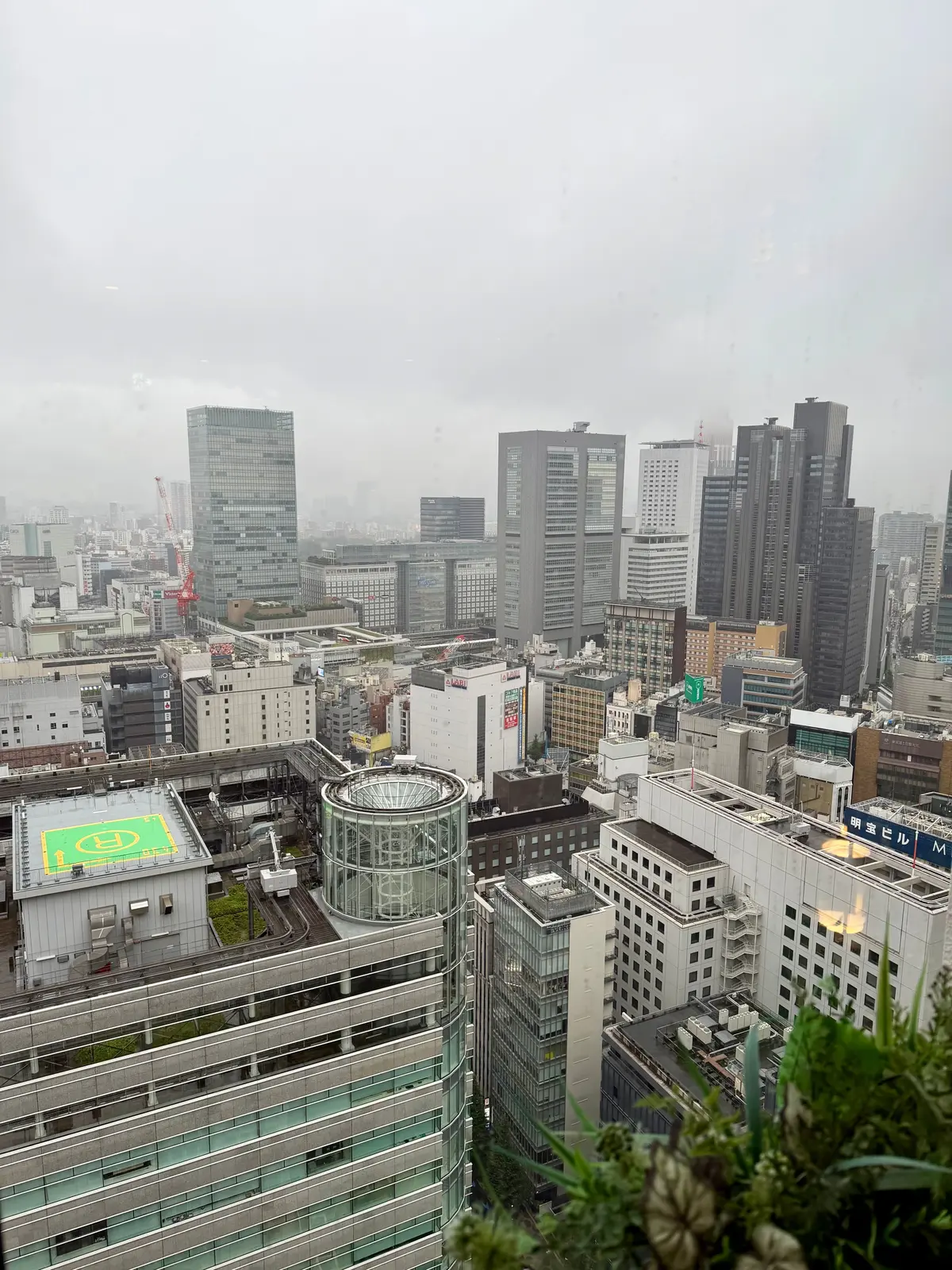 高層ビルが立ち並ぶ都市の風景。窓から撮影され、雨が降る曇天。ヘリポートのある建物や、ガラス張りの近代的なビル群も見られる。