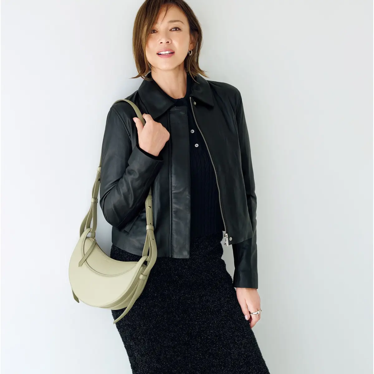 黒い襟付きセーターに黒のレザーJKT、黒の膝丈スカートを着用した女性。右肩にライトグレーのショルダーバッグをかけている。背景は白。