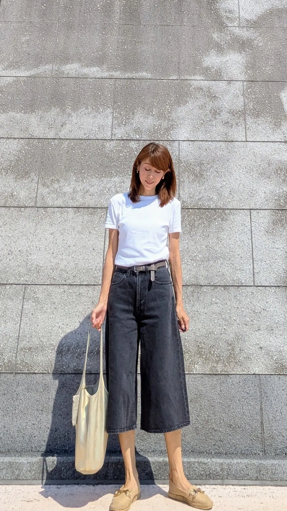 白いTシャツに黒いクロップド丈デニムを合わせた女性。ベージュのローファーとトートバッグを身につけ、グレーの壁の前に立っている。