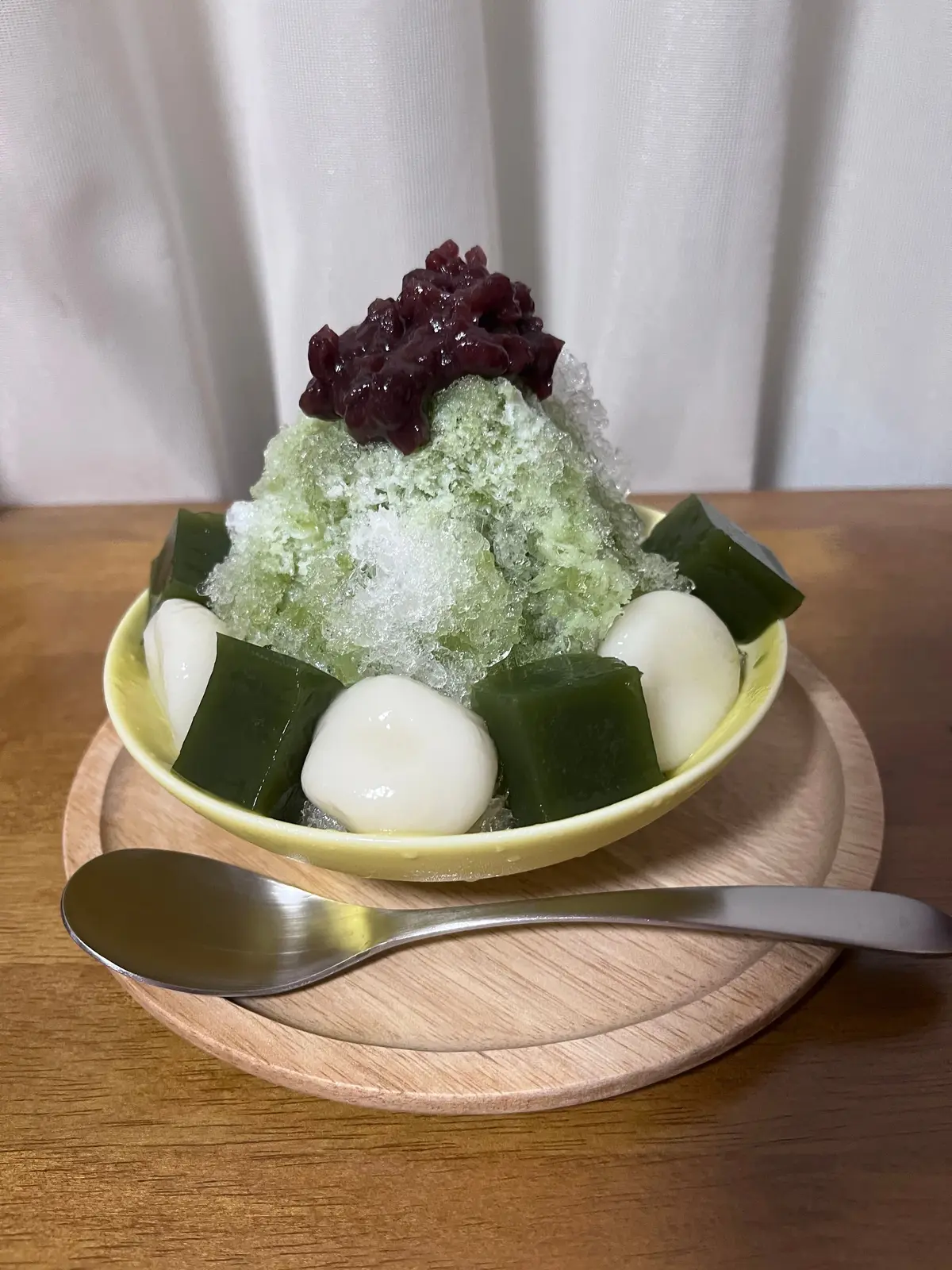 黄緑色の器に入ったかき氷。抹茶風味の氷の上に、紫色の豆の甘露煮と白玉、緑色の寒天が盛り付けられている。木製の皿とスプーンが添えられている。