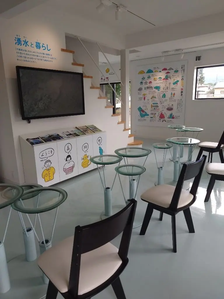 白を基調としたカフェの店内。丸いガラス天板と金属脚のテーブルが並び、木製の椅子が配置されている。左には案内板やイラストが飾られた棚、右には緑色のイラストが描かれた大きな掲示板がある。背景には窓から光が差し込み、木製の階段が見える。