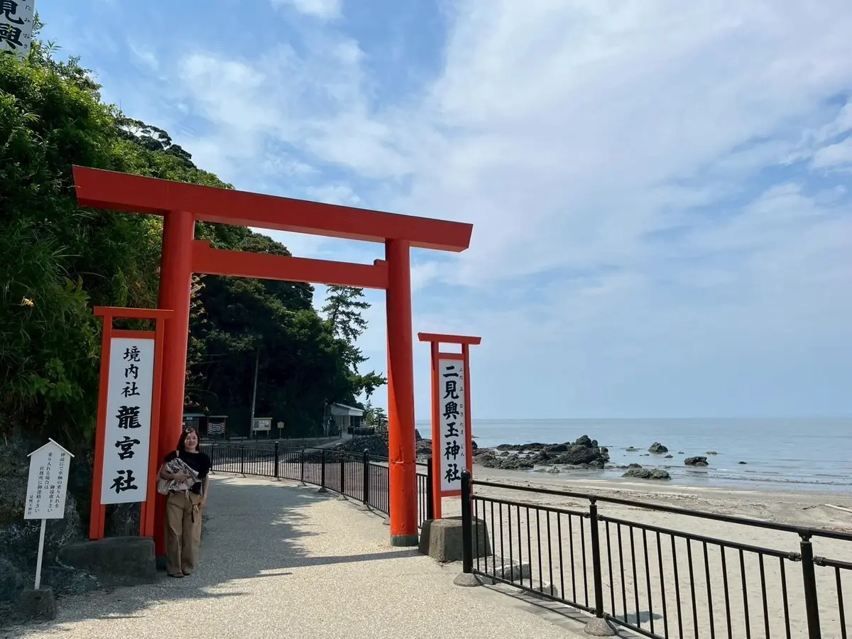海岸に面した鳥居の風景。鮮やかな朱色の鳥居が左右に立ち、中央には女性が立っている。背景には海と空が広がり、緑豊かな植栽も見られる。