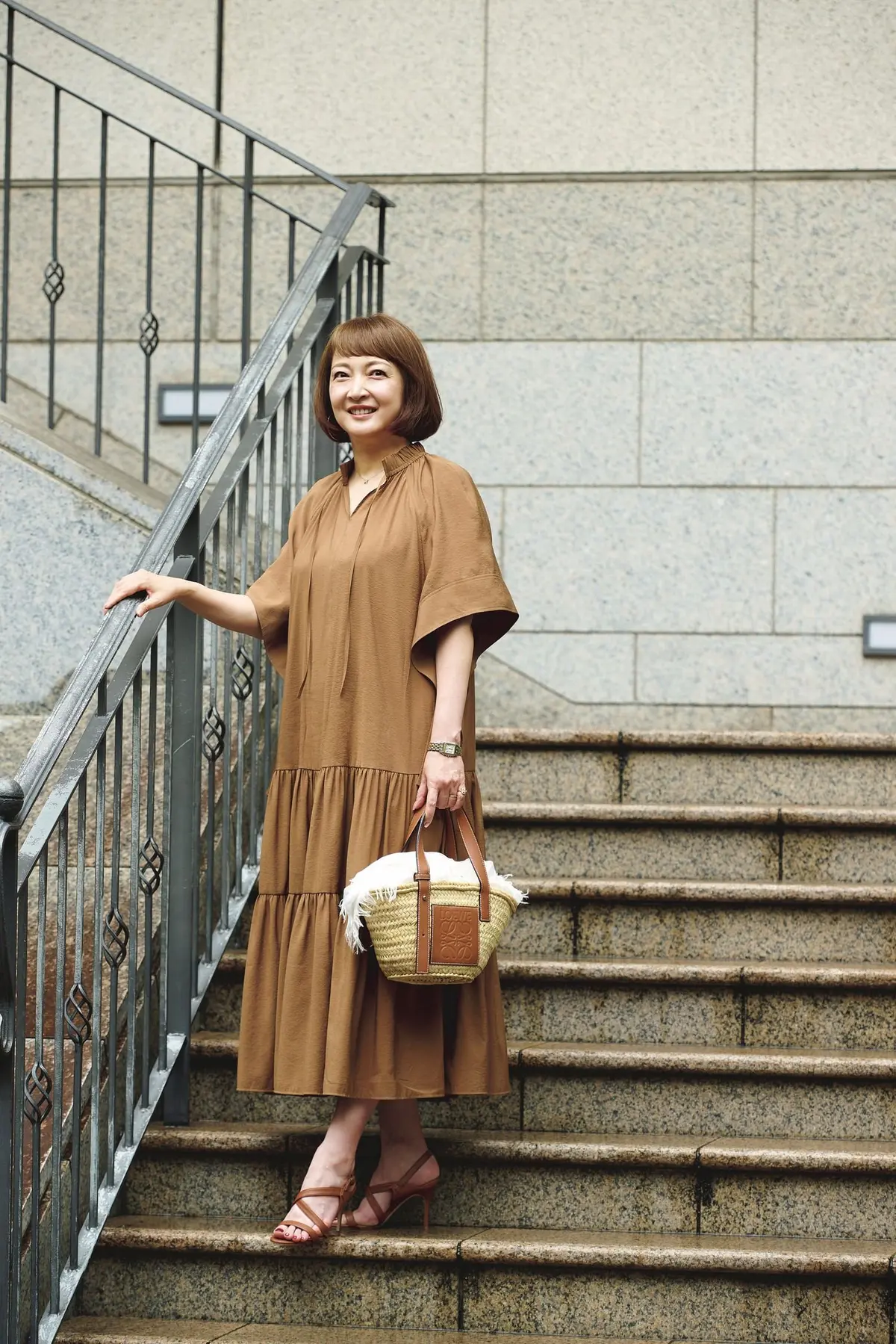 茶色のティアードワンピースを着た女性が、階段の手すりに手をつき、こちらを見ている。茶色のサンダルを履き、ロエヴェのバスケットバッグを持つ。背景はグレーの階段と壁。