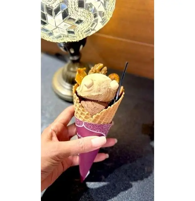 アイスクリームコーンにチョコレートとマシュマロのデコレーションがのったアイスクリームを手に持つ人。背景に模様のあるランプが設置されている。
