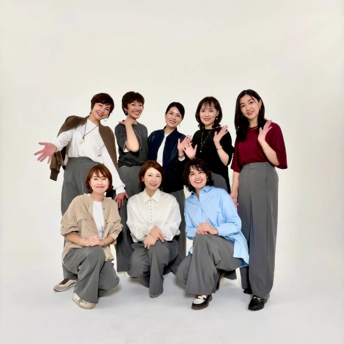 白背景をバックに、8人の女性が座ったり立ったりして写っているポートレート写真。全員がグレー系のシンプルな服装で、笑顔を見せている。手で挨拶やポーズをしている人もいる。