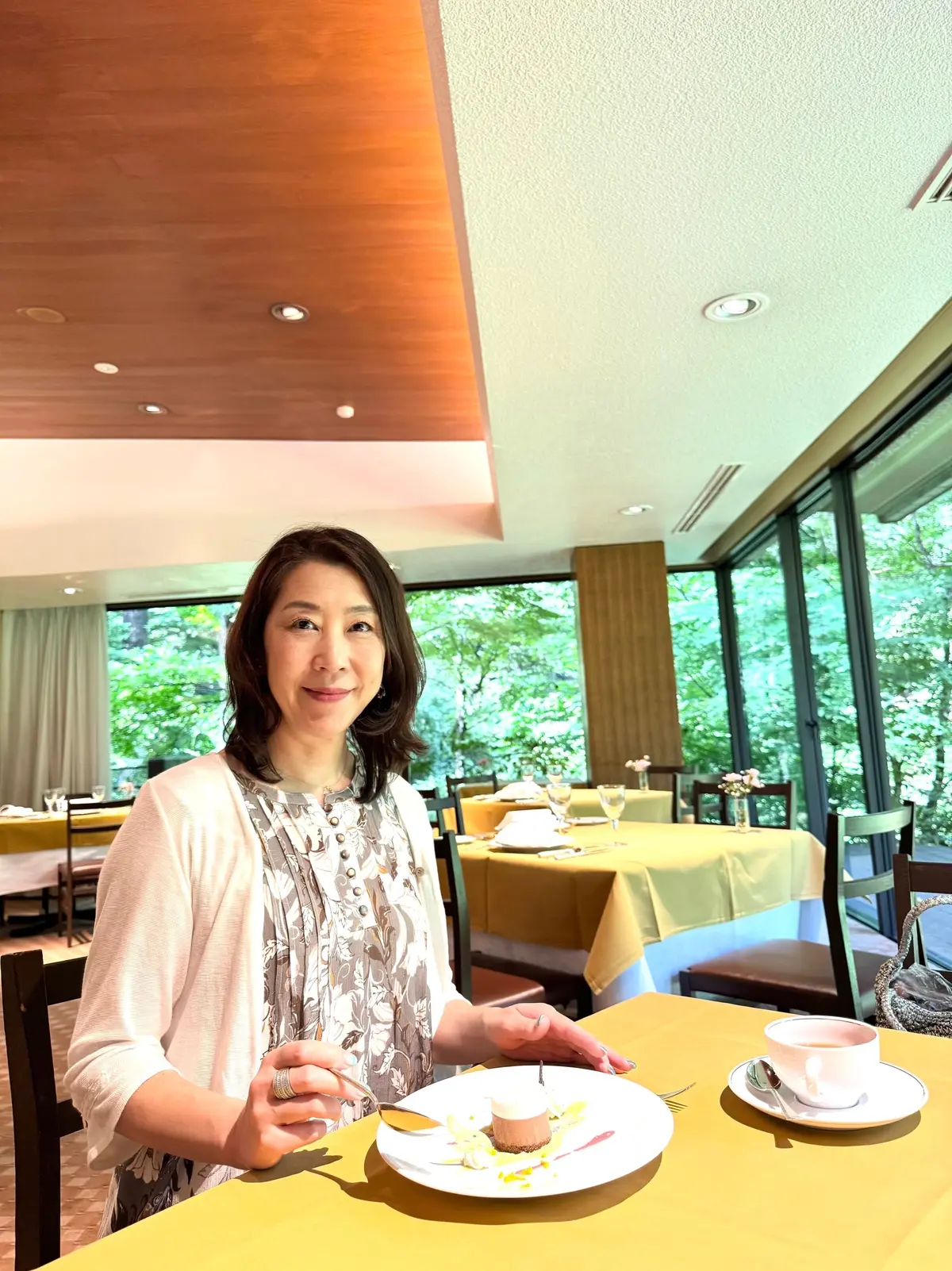 レストランのテーブルに座り、デザートを前に微笑む女性。白いカーディガンを羽織り、黄色のテーブルクロスが目立つ。背景には緑豊かな景色が見える。