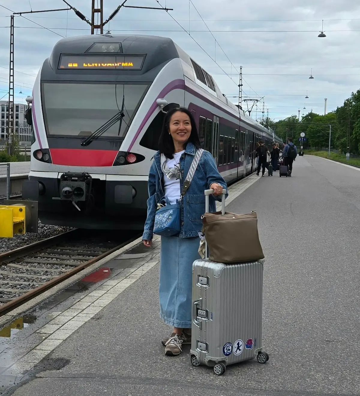 駅プラットフォームに立っている女性が、銀色のスーツケースと茶色の手荷物を持ち、電車が到着するのを待っている。電車は紫と白のデザインで、頭上に「LINHA VAMA」と表示されている。背景には木々や建物が見える。