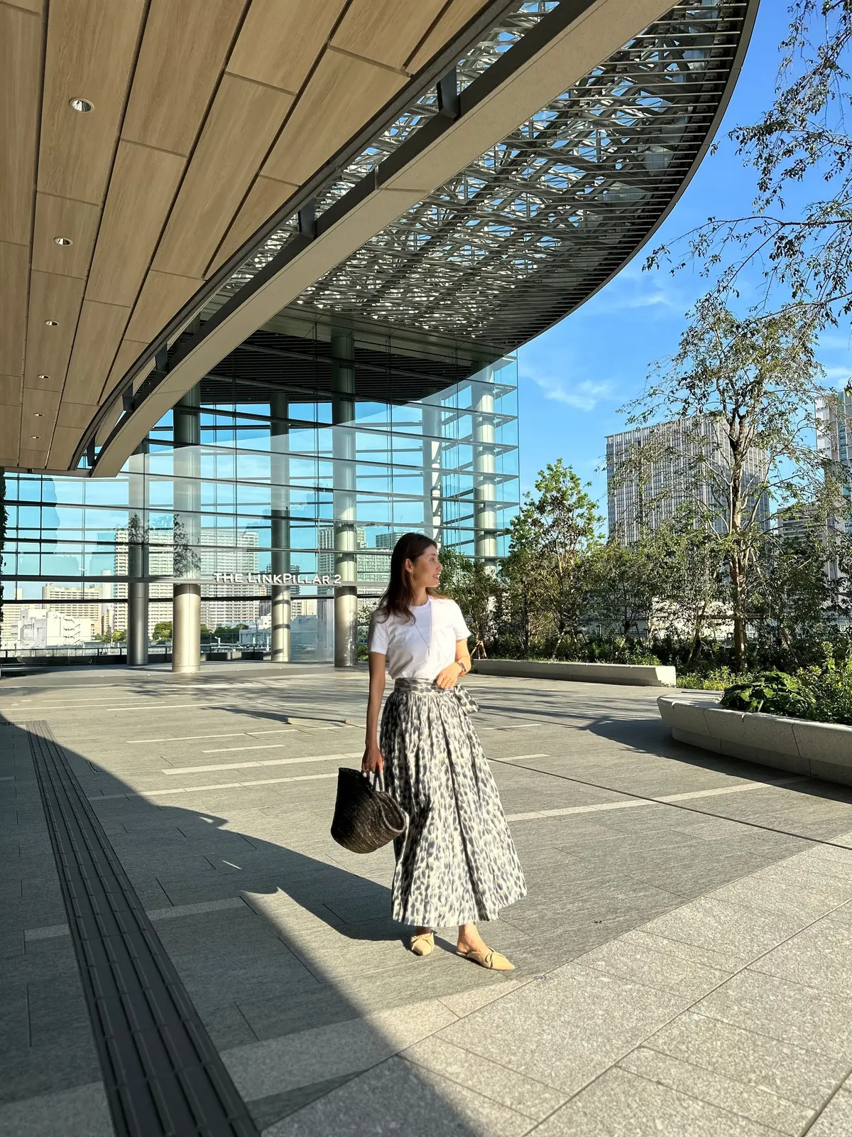 女性がレオパード柄スカートと白いトップスで、大きなトートバッグを手に歩いている。背景にはモダンなガラスの建物と木々が並ぶ都市空間。