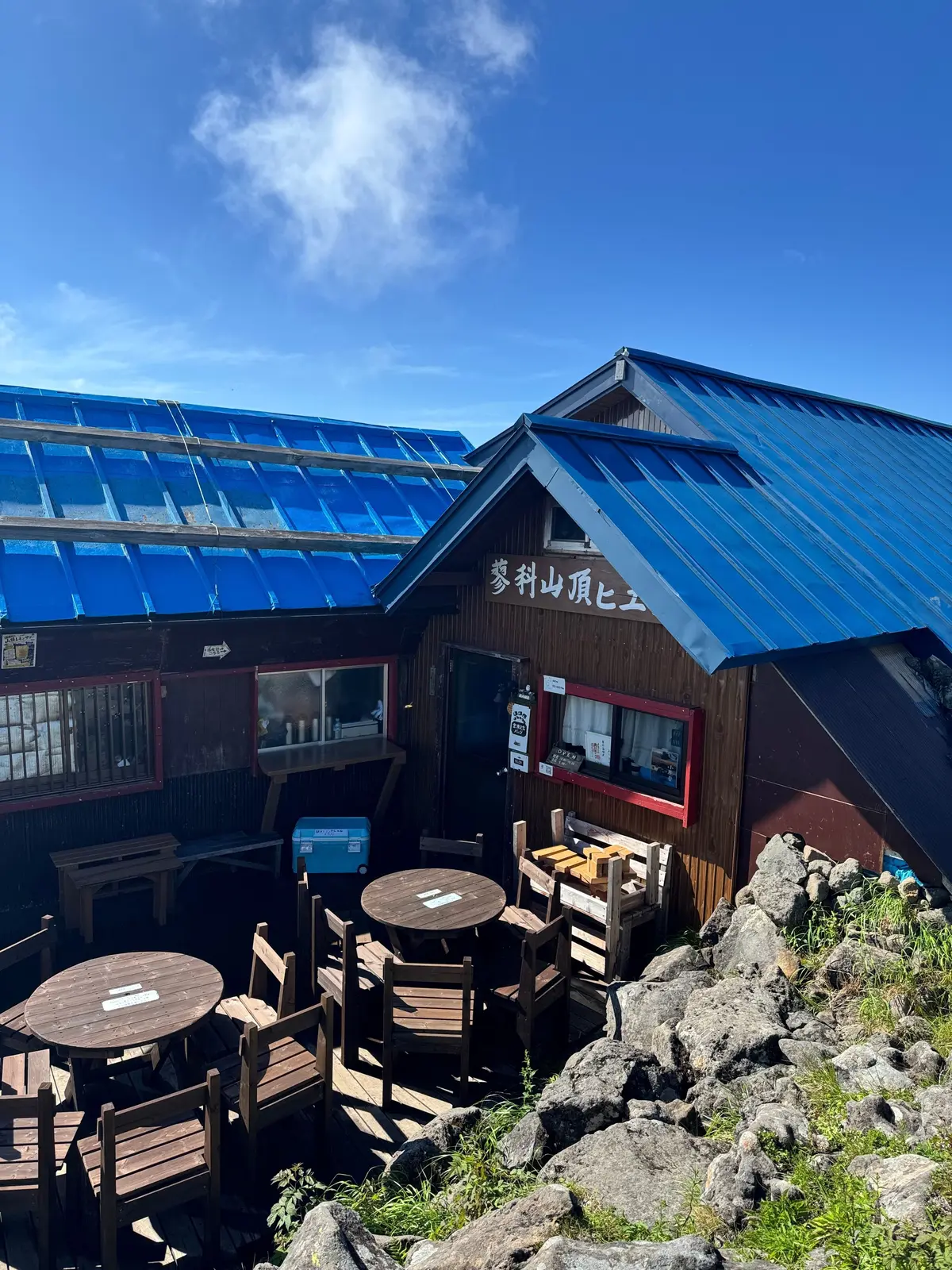 青い屋根の山小屋と、周囲の岩場、木製のテーブルと椅子が並ぶ風景。晴天の空には白い雲が浮かんでいる。
