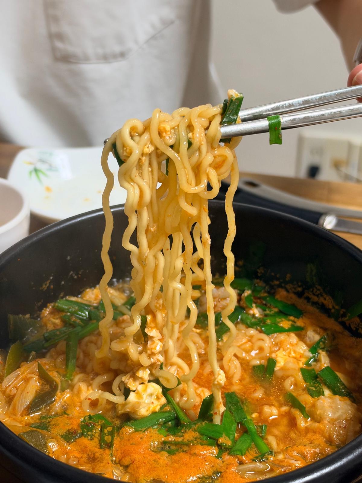 黒い鍋に入ったラーメンの麺が箸で持ち上げられている。スープは赤みを帯び、ネギや具材が浮かんでいる。背景には白いエプロンを着た人物と食器がぼやけて見える。