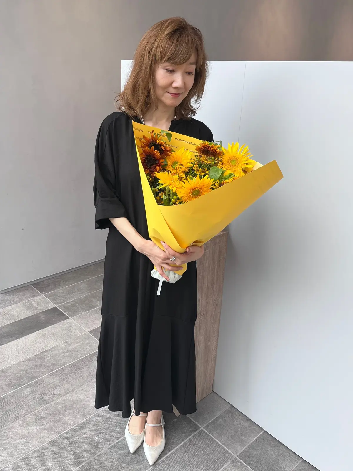 黒いティアードワンピースを着た女性が、黄色いひまわり束を持つ。浅い栗色の髪を肩まで伸ばし、控えめな微笑みを浮かべている。背景はベージュ色の壁と床。