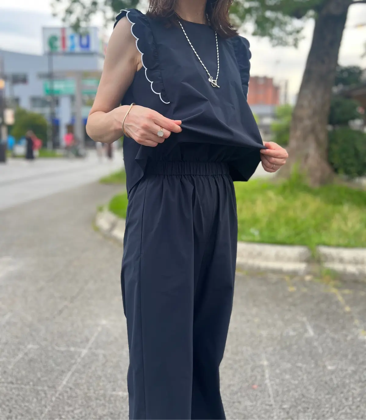 黒いフリル付きノースリーブトップスと、ウエストがギャザーになった黒いパンツを着用した女性。背景には道路と緑の木々が見える。シンプルなシルバーのネックレスとリングを身に着けている。
