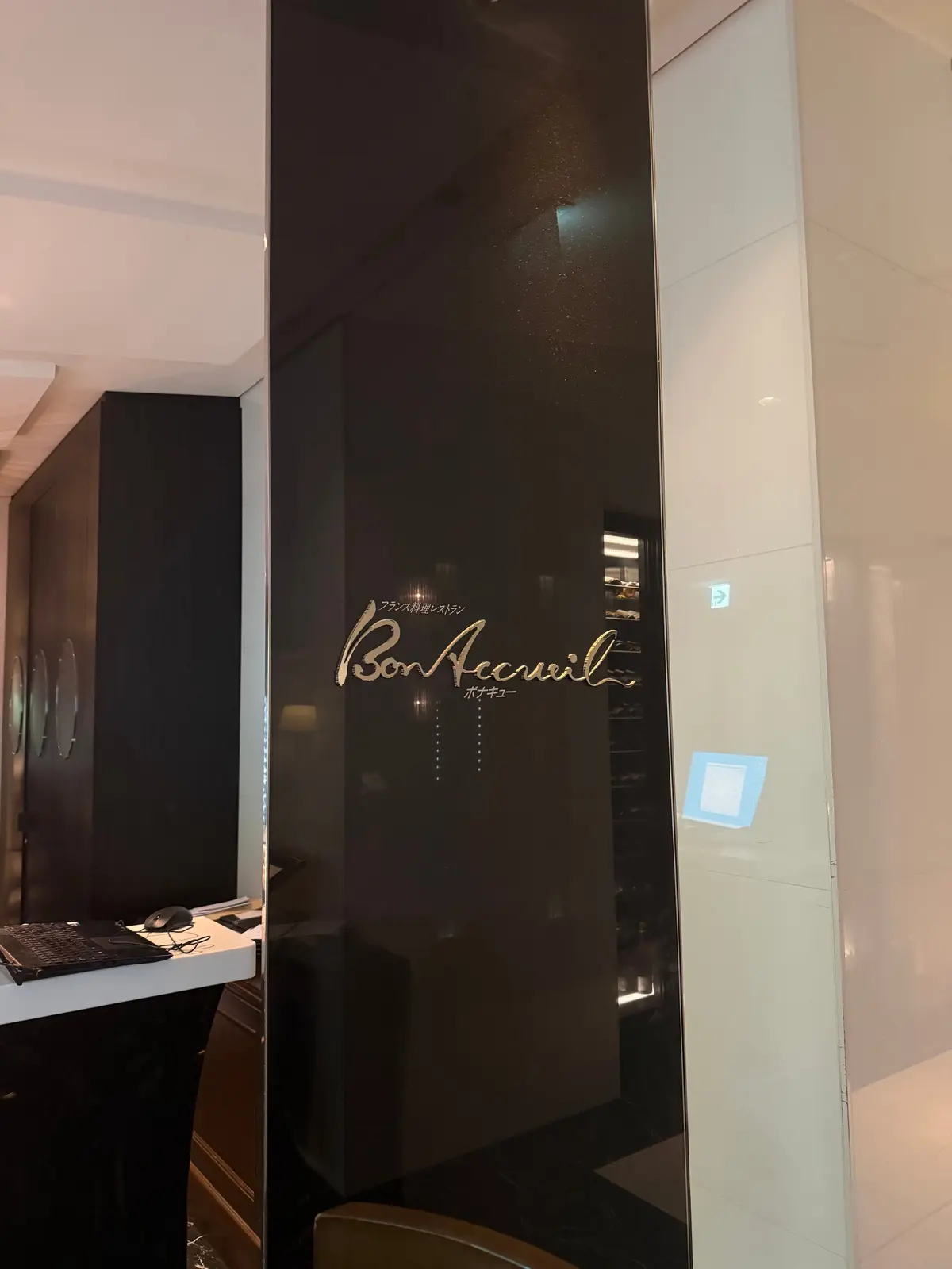 黒い柱に金色の手書き風文字で「Bon Accueil（ボン アクル）」と表記された看板。右には白い壁とドア、奥にはエレベーターが見える。