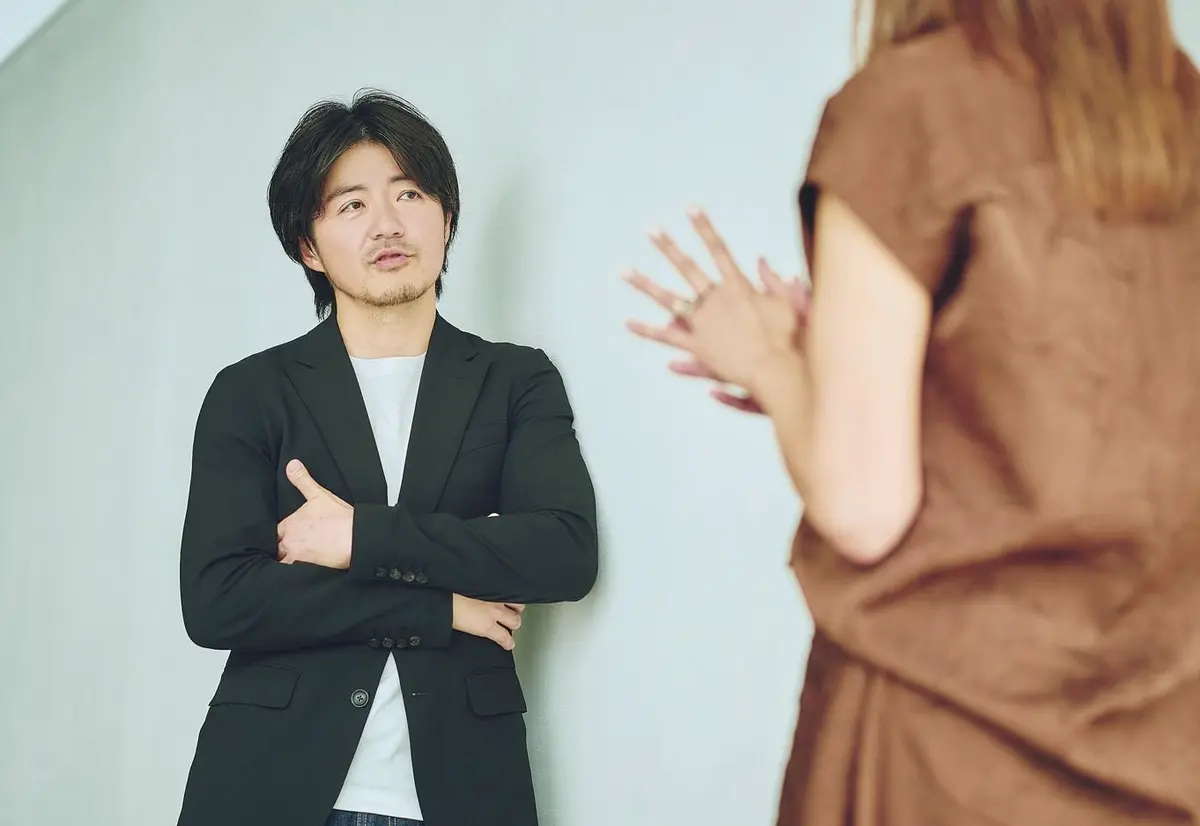 黒いジャケットを着た男性が腕を組み、壁に寄りかかっている。背景には茶色の服を着た女性が立っており、両手を広げている。